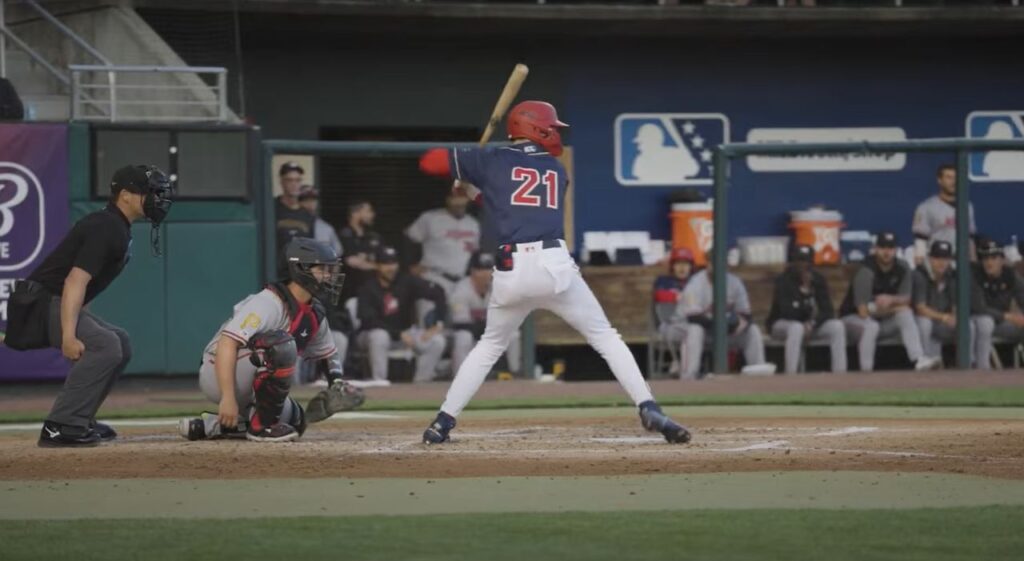 Back of baseball player at bat, wearing dark blue jersey with red #21 and white pants, holding baseball bat, with catcher squatting on left side.