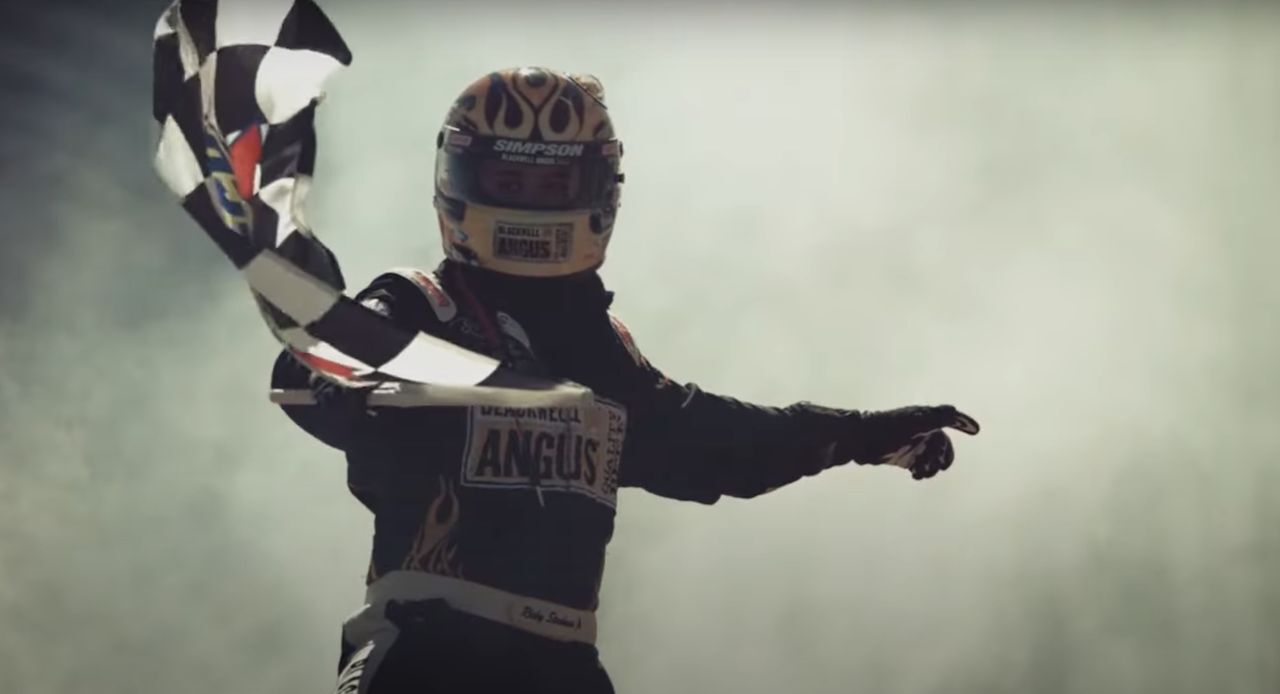 Person wearing race car driver outfit with helmet waving checkered flag with smokey background