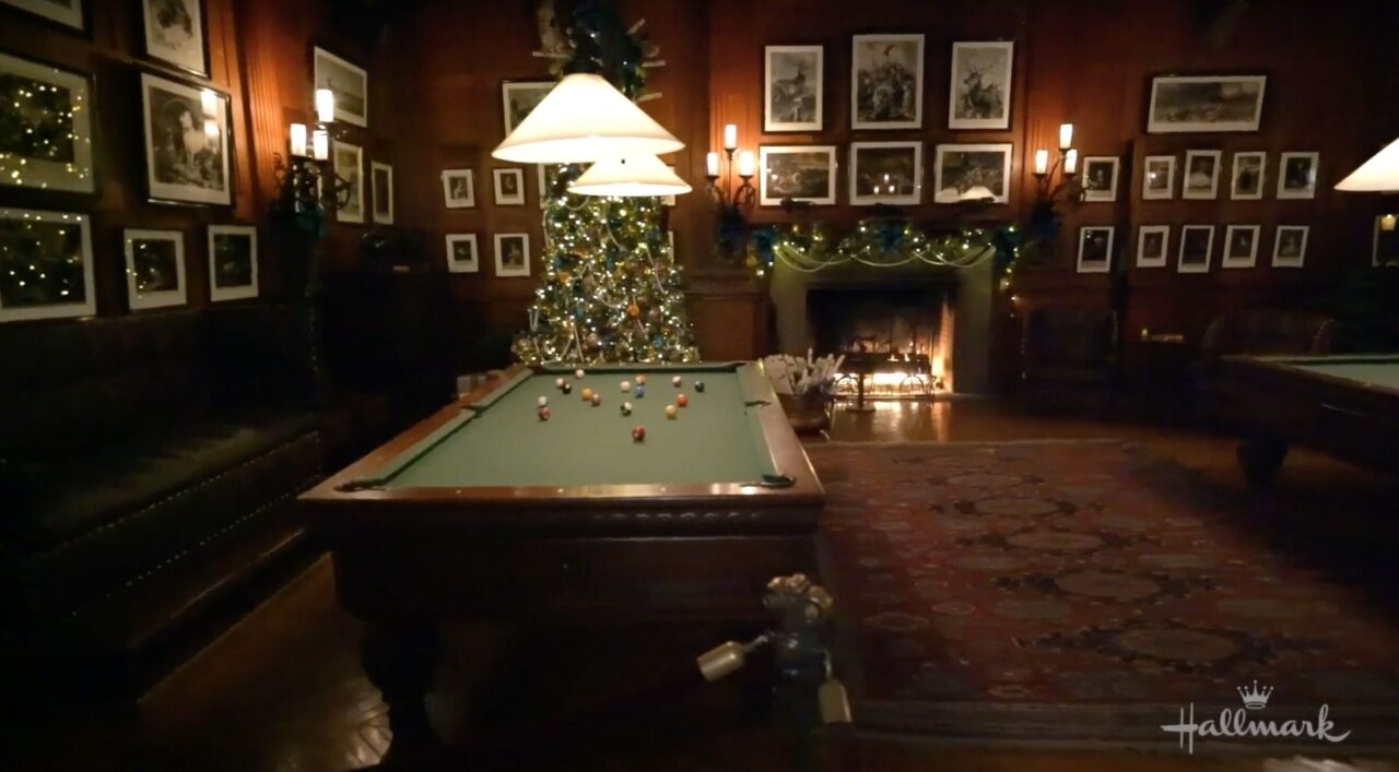 A billiards table and balls with two pendant lights in a room with multiple framed pictures on the walls, a fire in the fireplace and a Christmas tree with lights and decorations