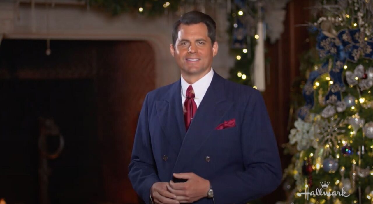 Male actor from "A Biltmore Christmas" wearing navy blue suit and burgundy tie and pocket square, standing near Christmas tree.